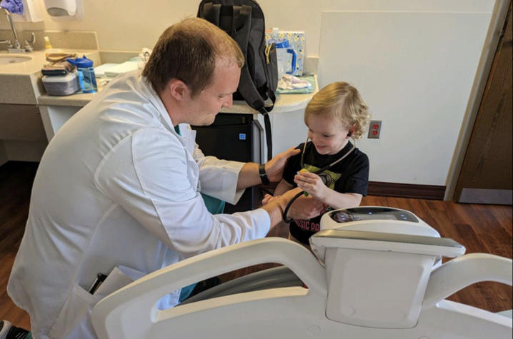 Andre Arnold, dressed in his white coat, helps a child hear their own heart beat using his stethoscope.