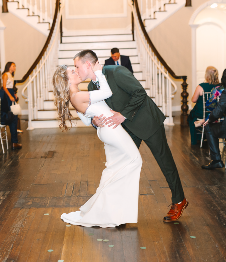 A bride in a white gown and a groom in a green suit share a kiss in a ballroom.