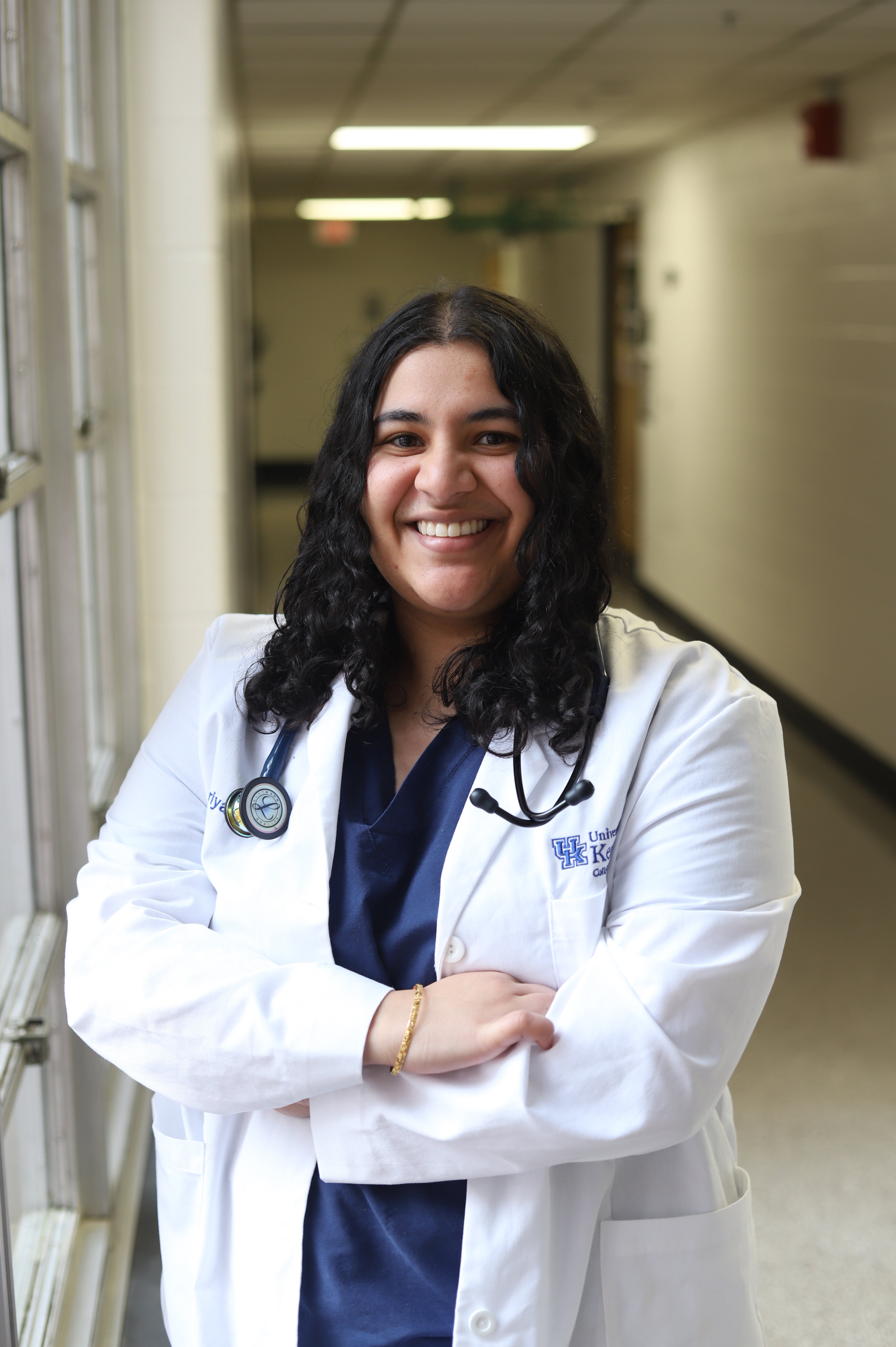 Supriya Challa stands with arms crossed in hallway, smiling, wearing a white coat