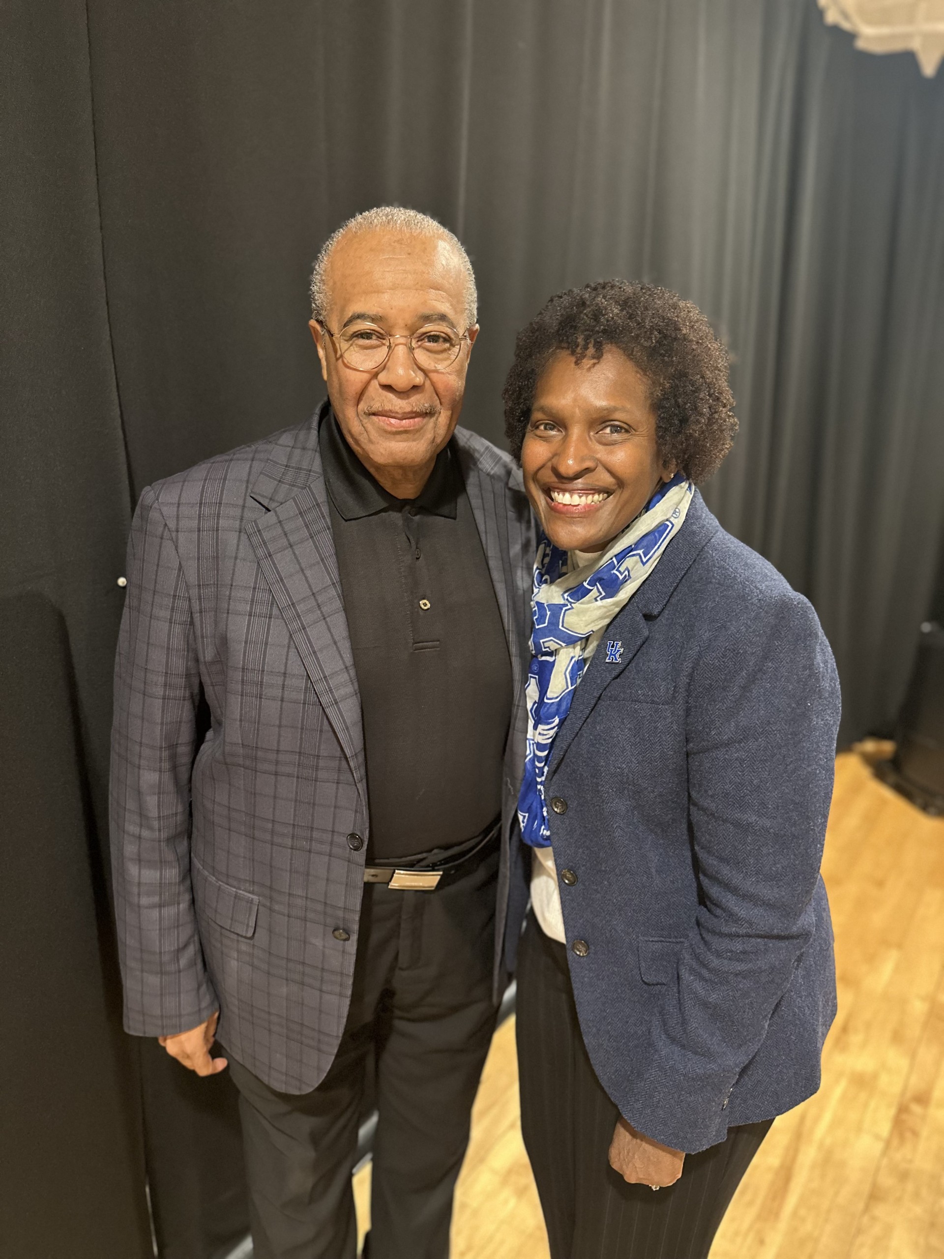 Dr. Lovoria Williams and Pastor Joseph Owen pose together, smiling warmly. Dr. Williams wears a navy blazer with a blue and white scarf, while Pastor Owen wears glasses, a dark polo shirt, and a gray plaid blazer. They stand in front of a black curtain in an indoor venue.