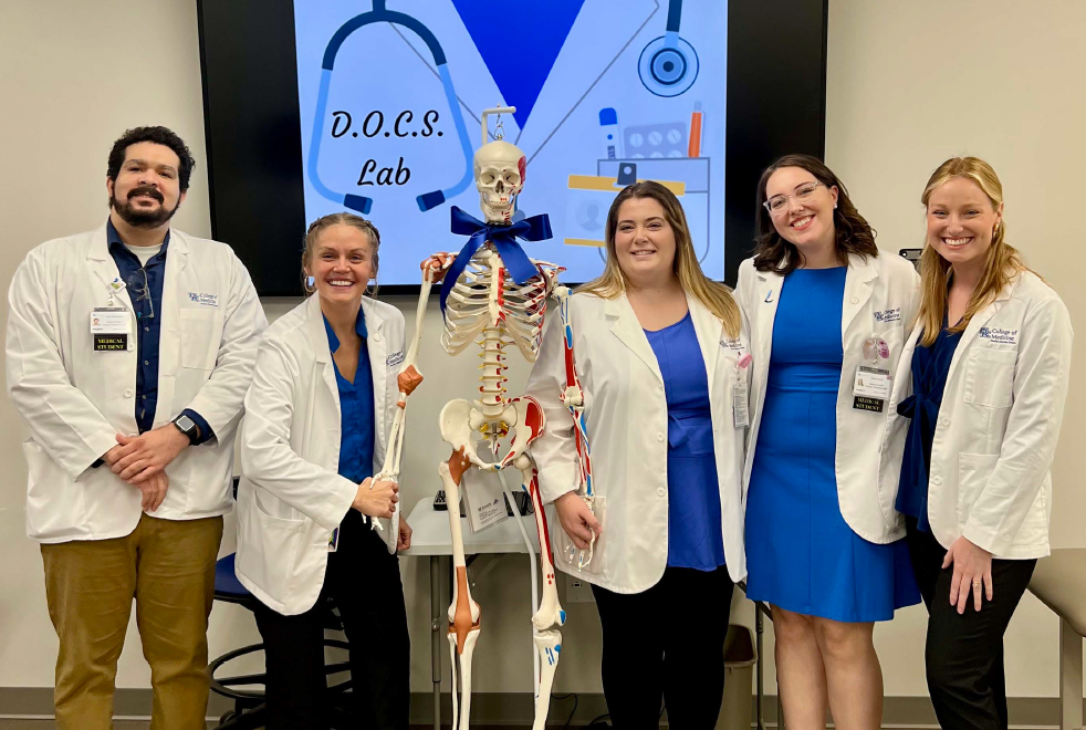 Five people in lab coats and blue attire stand smiling beside a skeleton model wearing a bow tie. A sign behind reads "D.O.C.S. Lab."