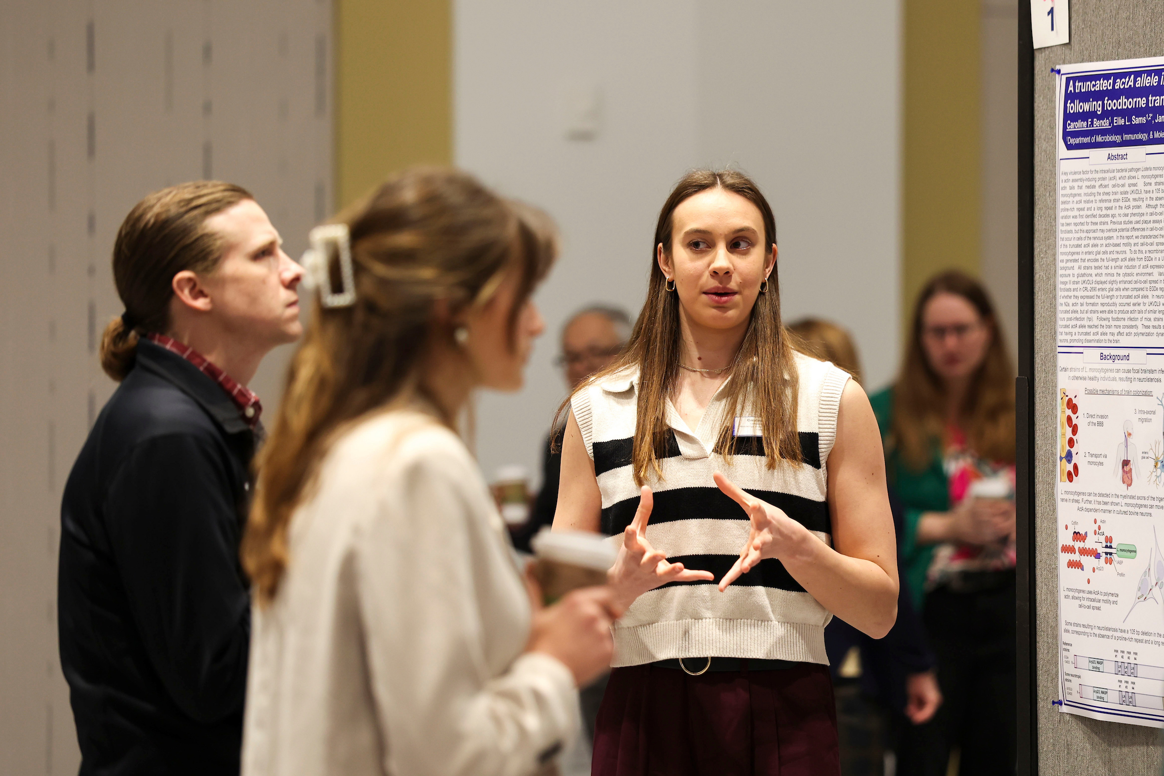 Presenter discusses her research poster with attendees during a poster session, gesturing as others listen and view the display.