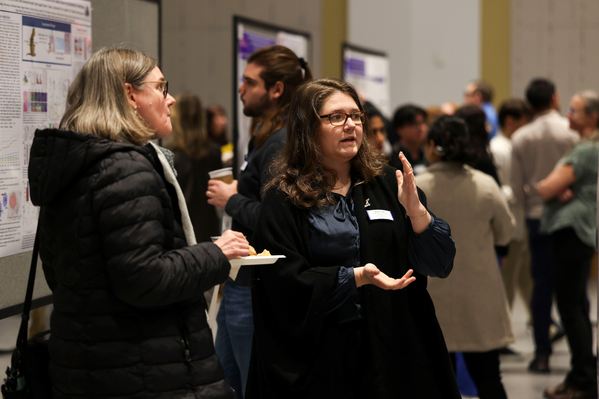 Two attendees converse beside research posters during a busy poster session, with other guests mingling in the background.