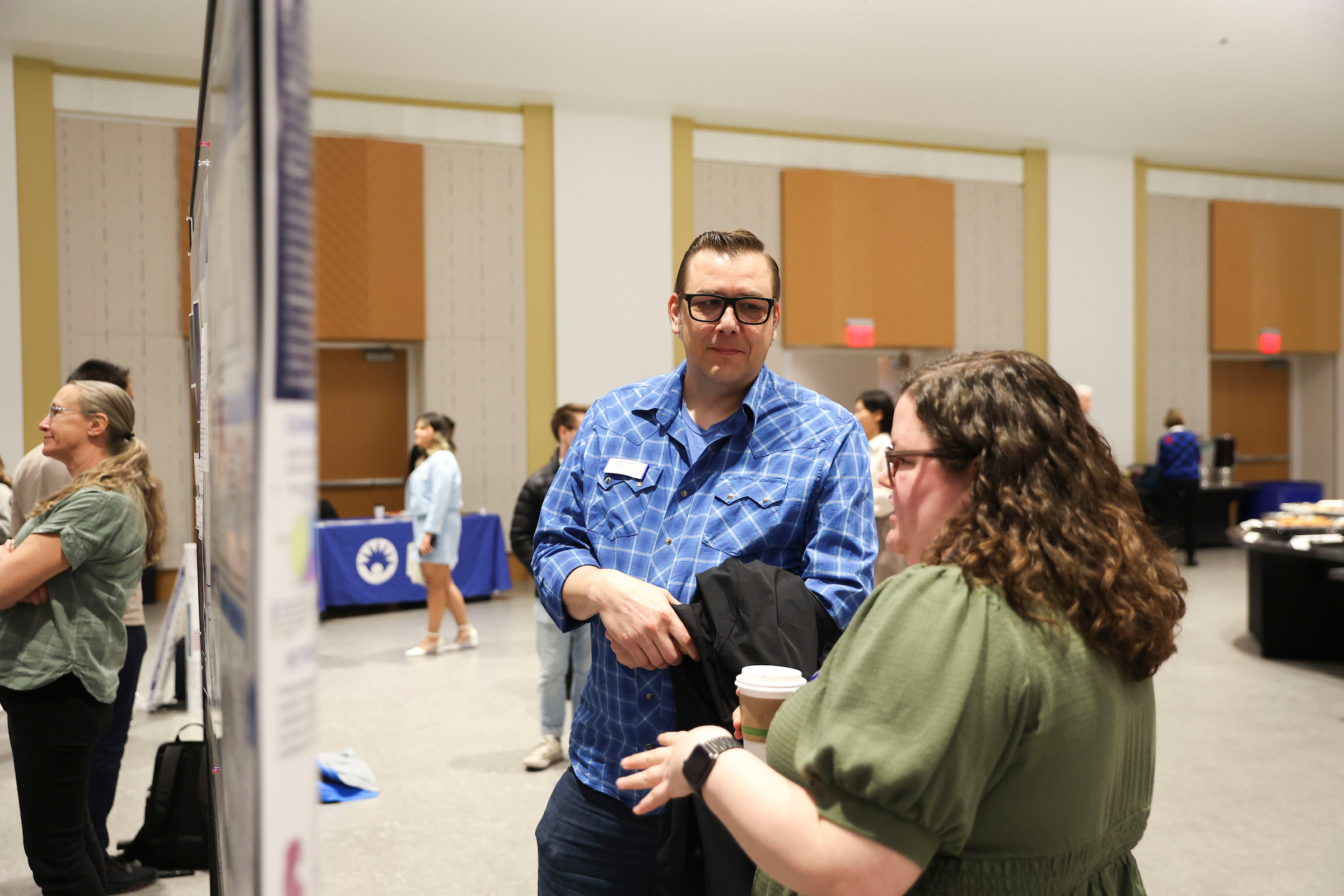 Two attendees discuss a research poster during a poster session, with other guests and poster displays visible in the background.
