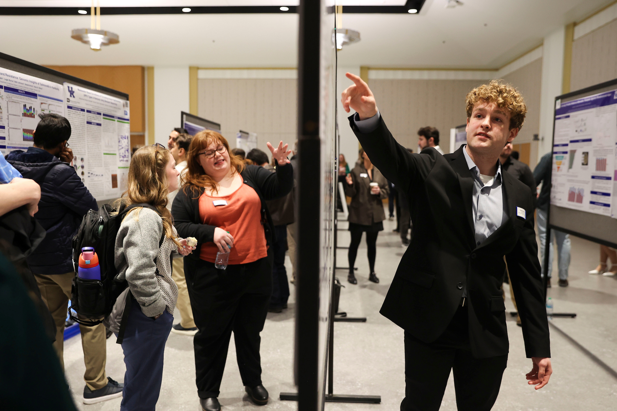 Presenter gestures toward a research poster while explaining findings during a busy poster session with attendees and displays in the background.