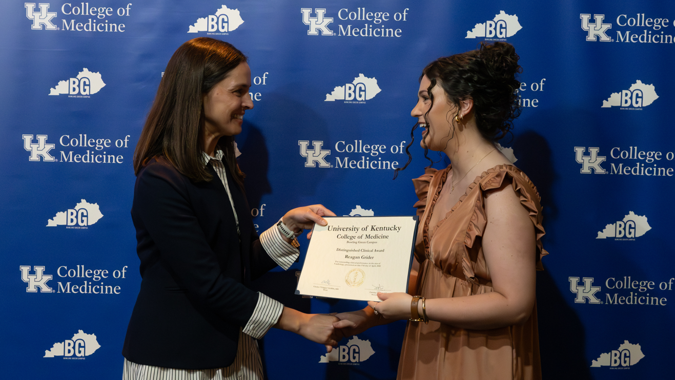 Two smiling individuals exchange a certificate in front of a University of Kentucky College of Medicine backdrop, conveying achievement and celebration.