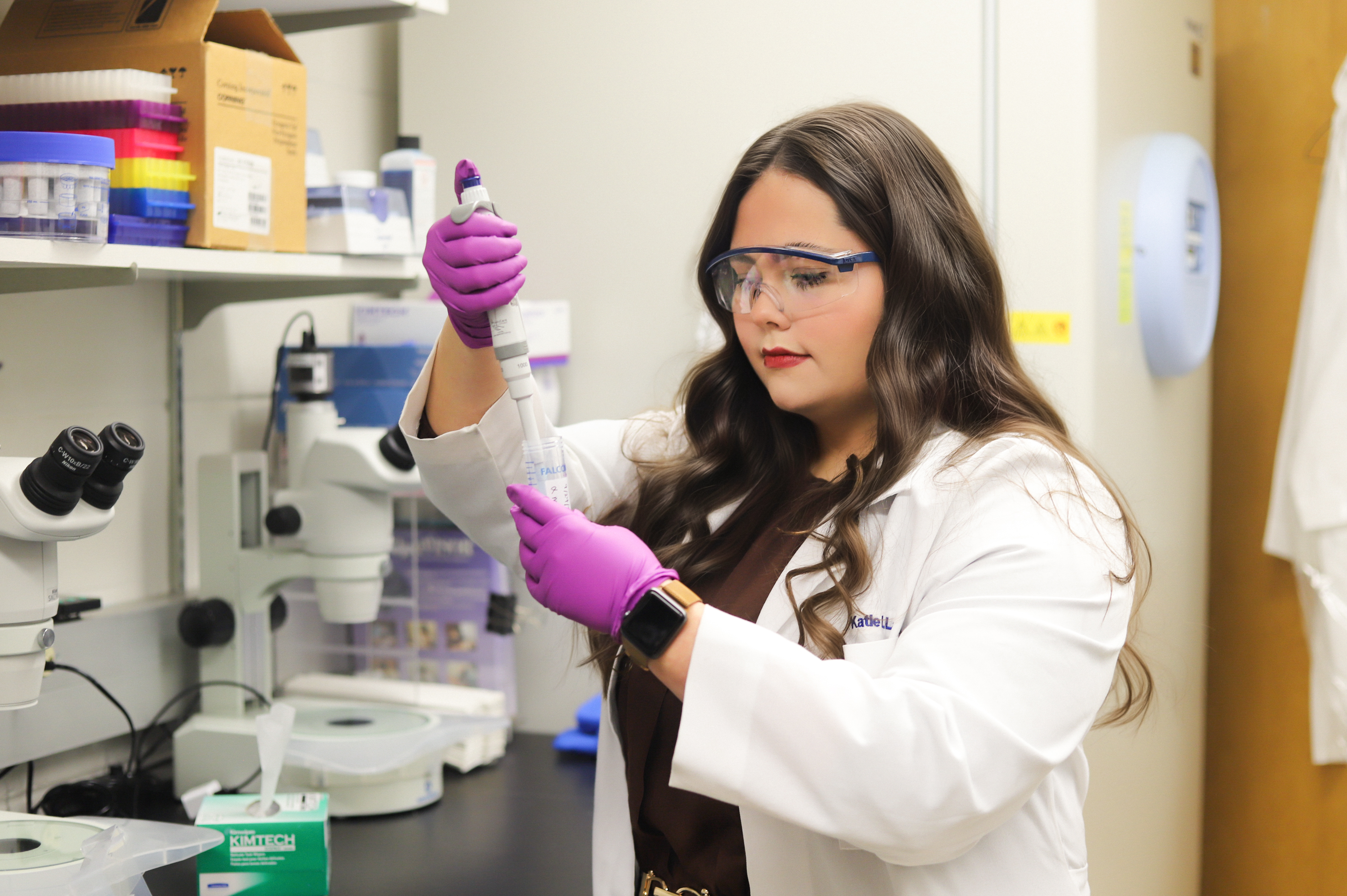 A scientist in a lab coat uses a pipette in a lab, wearing safety goggles and purple gloves. Shelves with lab equipment and microscopes are around her.
