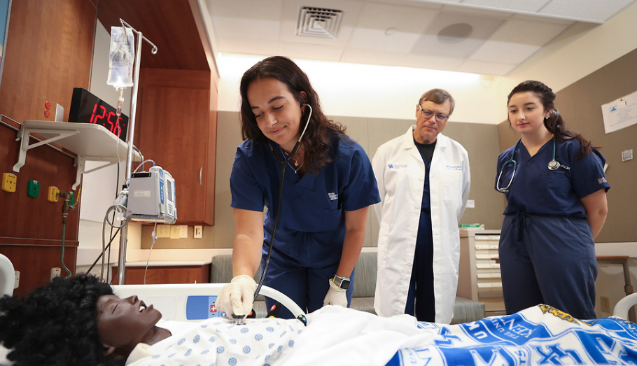 Medical simulation with two med students in blue scrubs and a white coat attending to a patient mannequin in a hospital room setting.