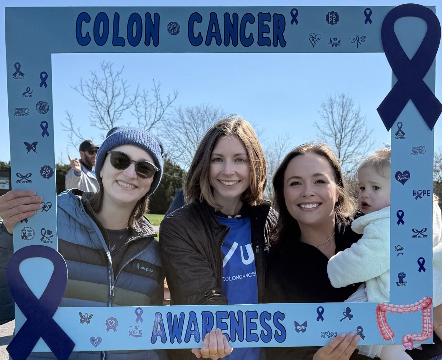 In a large cardboard frame that reads "Colon Cancer Awareness" three women smile. One holds a baby.