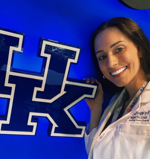 Alicia, in a white coat, poses next to a neon UK sign