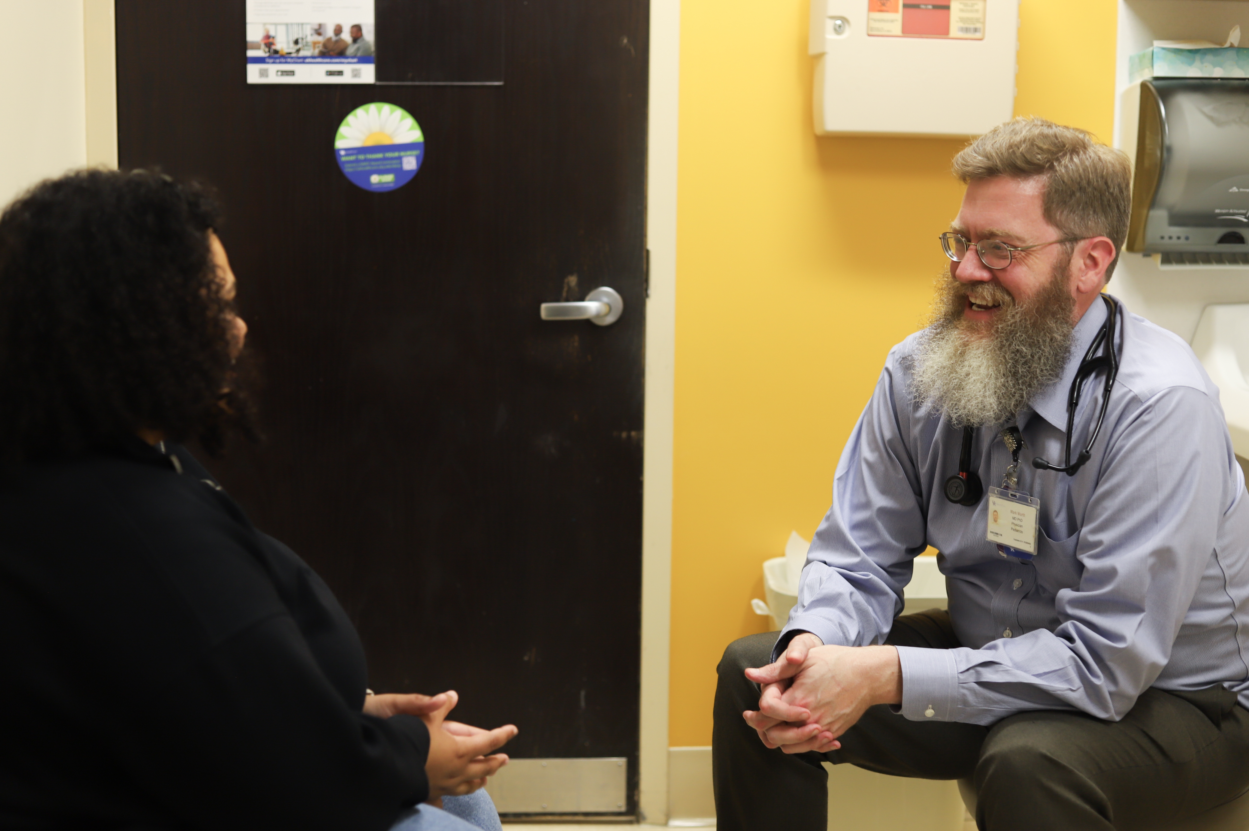 In a yellow exam room, Dr. Wurth sits to the right, across from a patient, smiling and talking. 