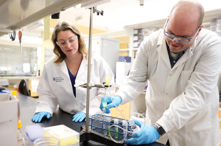 Two researchers wearing lab coats, safety glasses and gloves work together in a laboratory, preparing and transferring liquid samples into test tubes and vials on a lab bench.