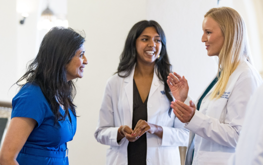 Three women stand talking. On the left in a blue dress, and in the center and right in a white coat. 