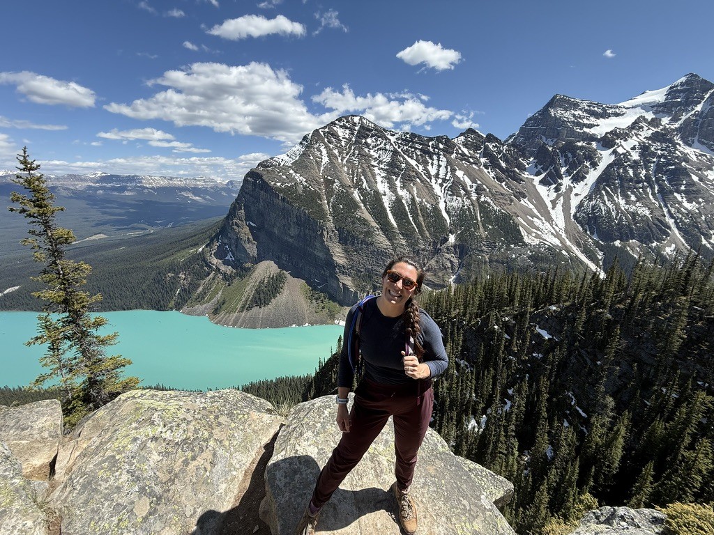 Maddie Mitcham standing at the top of a mountain with the view of mountains and a vibrant blue lake behind her. 