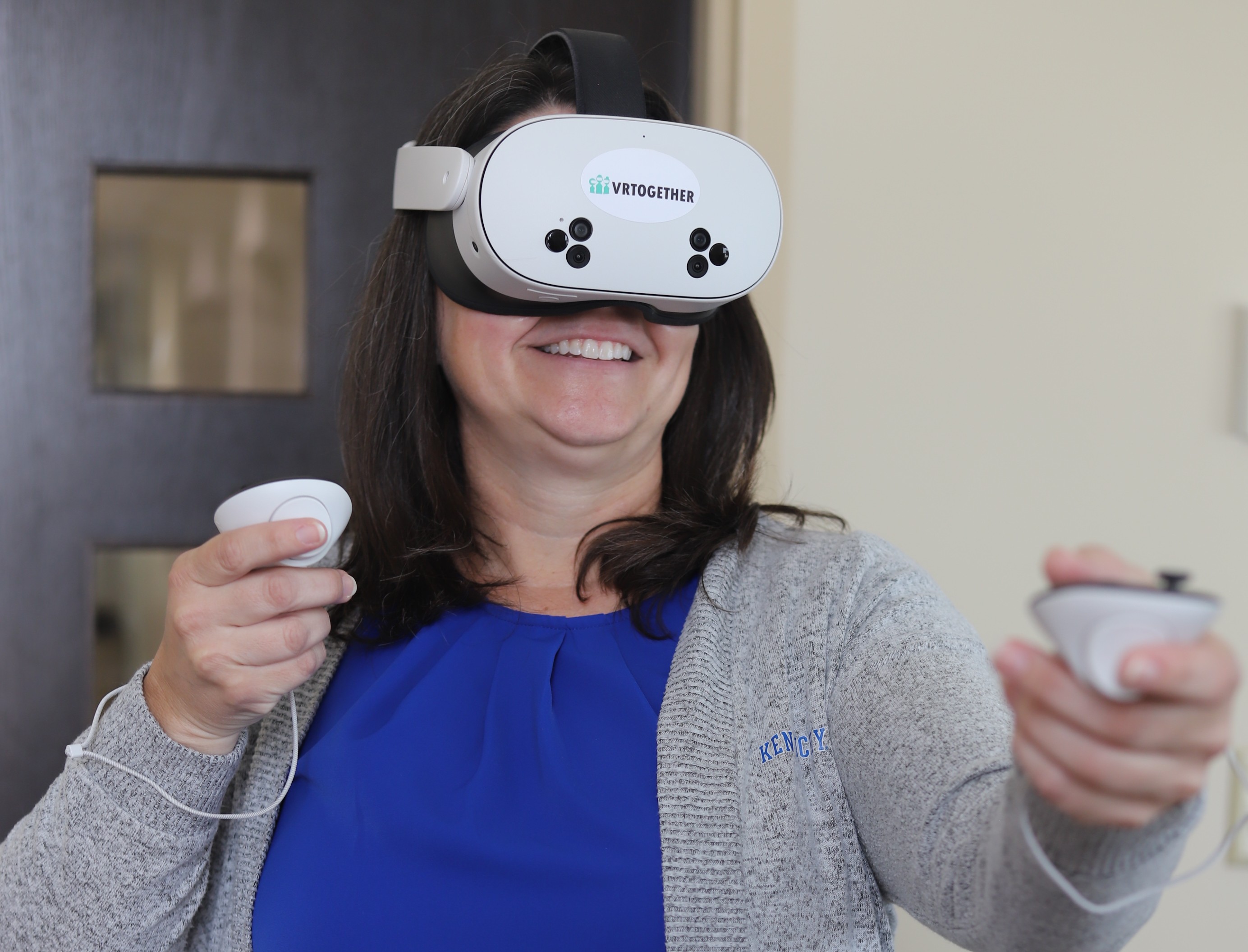 A women in a blue shirt and gray UK cardigan wears a VR headset labeled "VRTogether" and holds a white controller in each hand. 