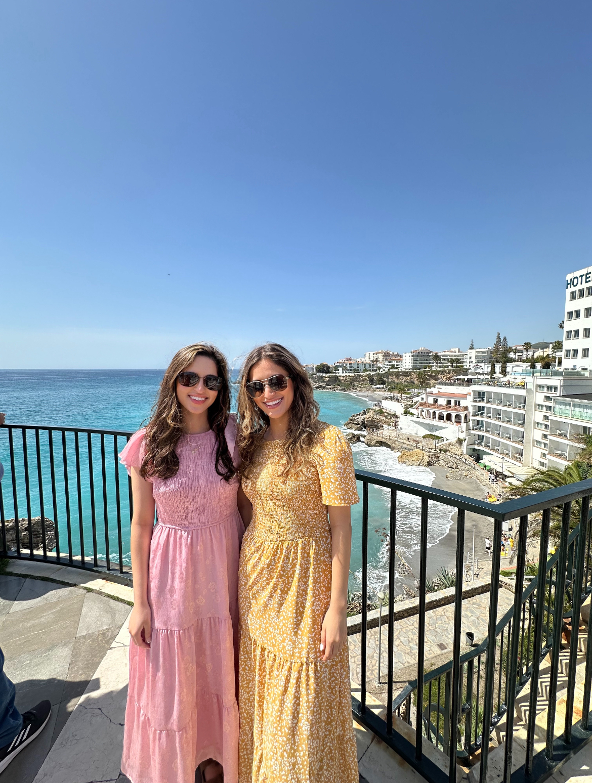 Aliya (left) standing with her twin sister (right) on a balcony with the ocean view behind them. 