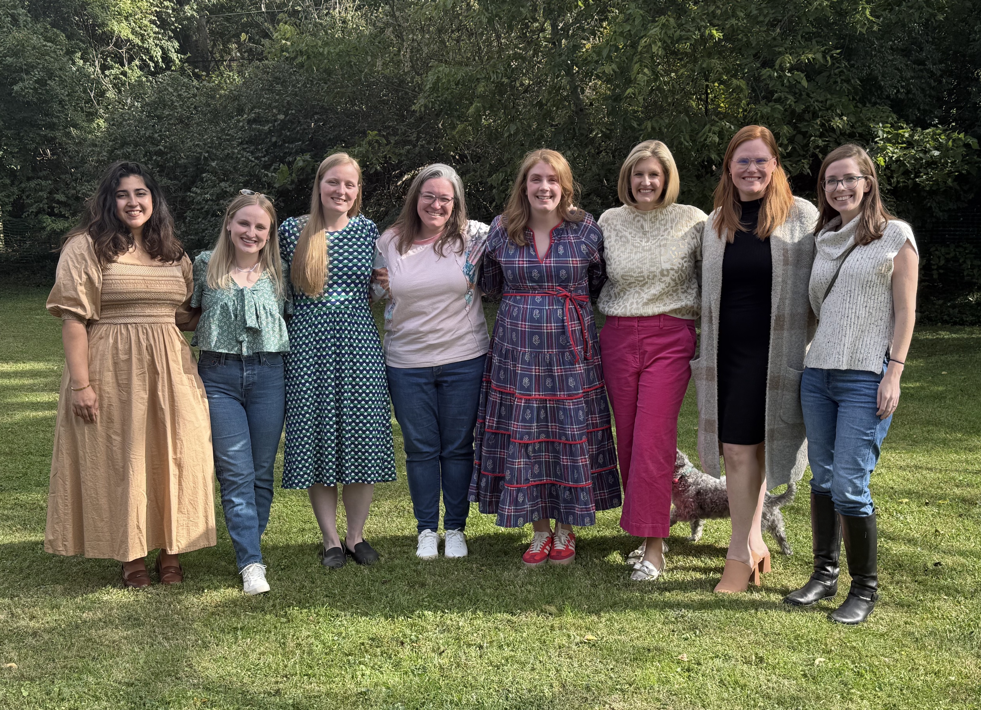A group of eight women in various outfits stand together outdoors on a grassy area surrounded by trees.