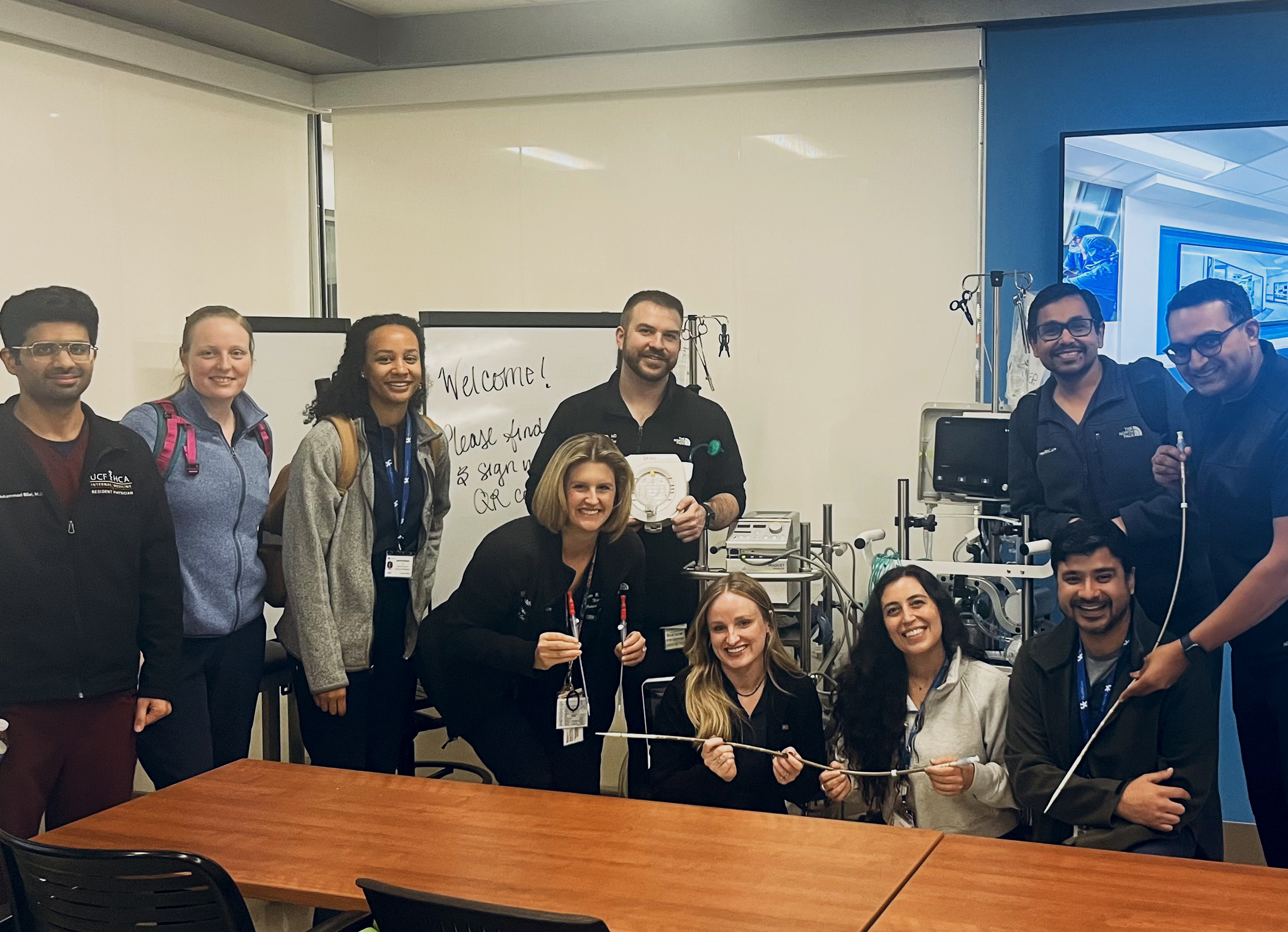 A group of healthcare professionals pose in a medical training room, holding equipment, with a whiteboard in the background reading "Welcome!" and various medical devices visible.