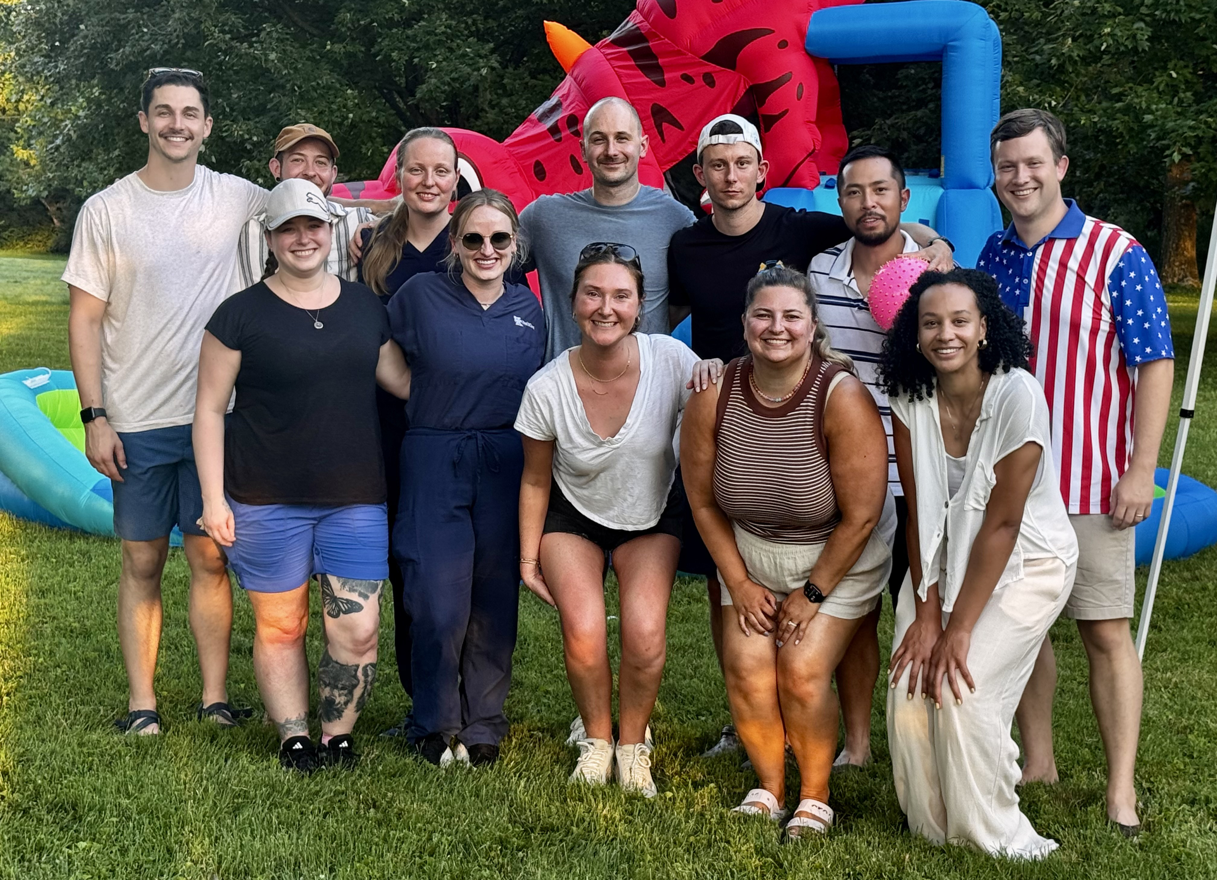 A diverse group of twelve people poses together on a grassy field with a colorful inflatable water slide in the background.