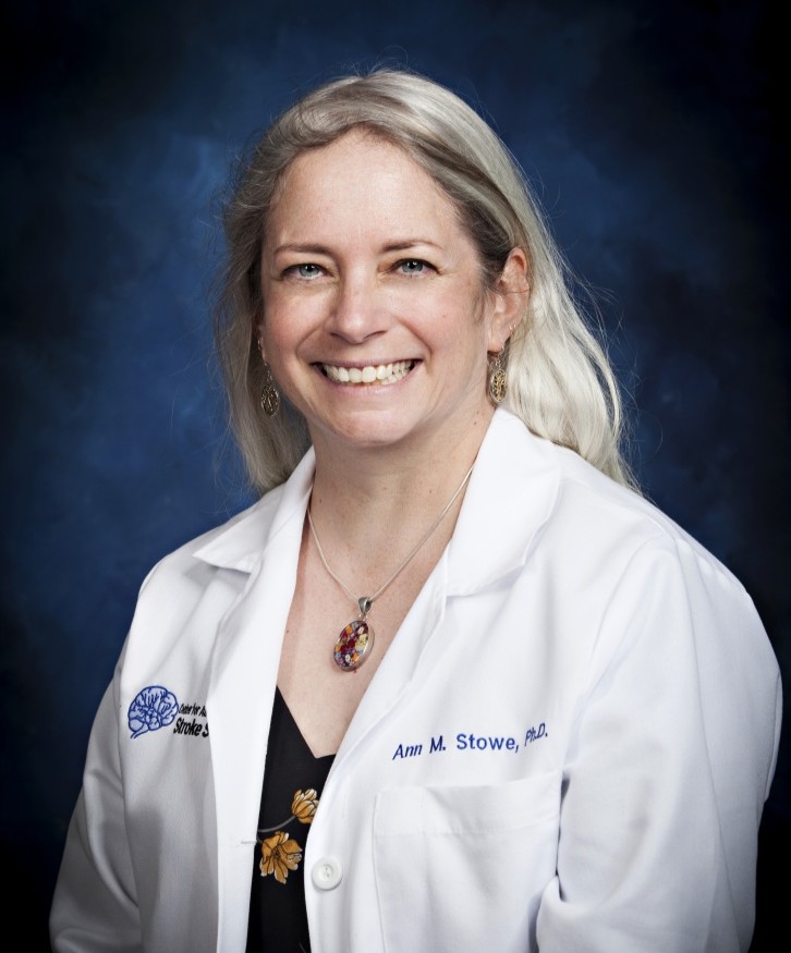 Ann Stowe headshot, a smiling blonde woman wears a white coat in front of cloudy dark blue background