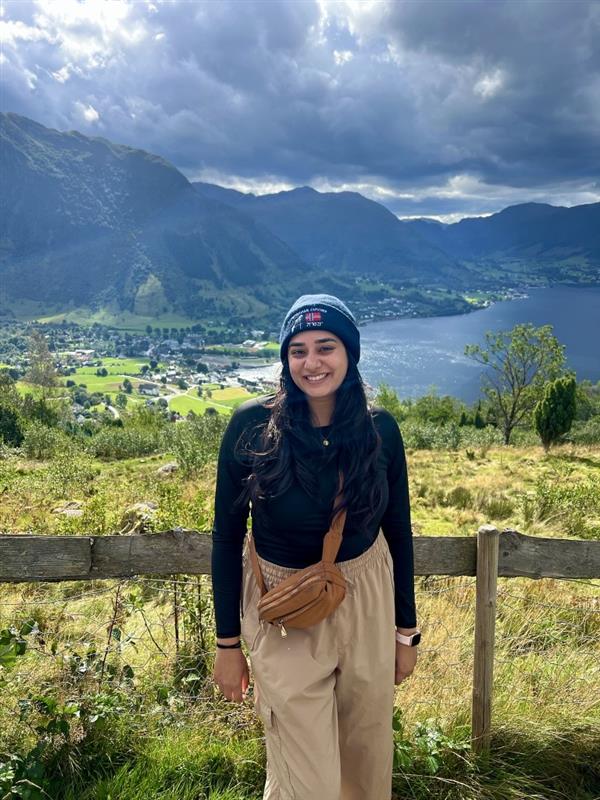 Tulsi smiling in a picture of her hiking with a beautiful grassland behind her with the ocean and mountains