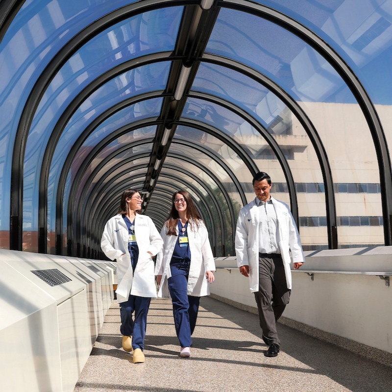 Two women in white coats and blue scrubs and one man in a white coat, white shirt, and brown pants walking side by side in a well-lit pedestrian tunnel