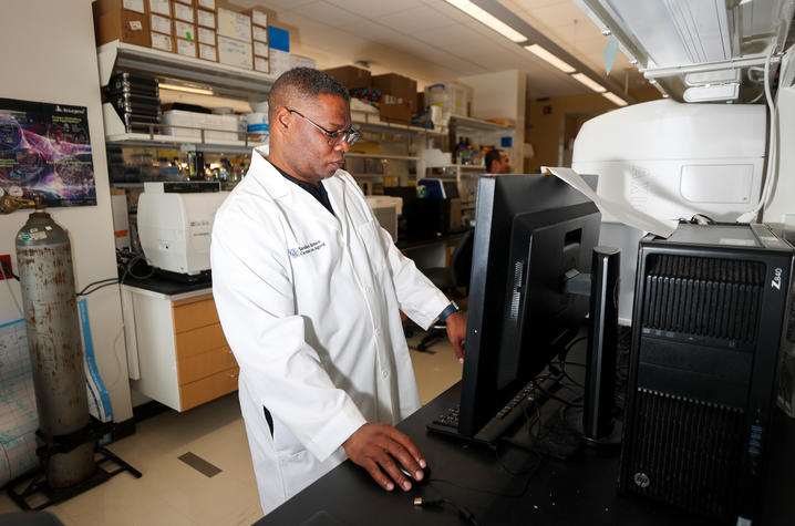 Daniel Lee, wearing a white lab coat, works at a computer beside research equipment in a laboratory.