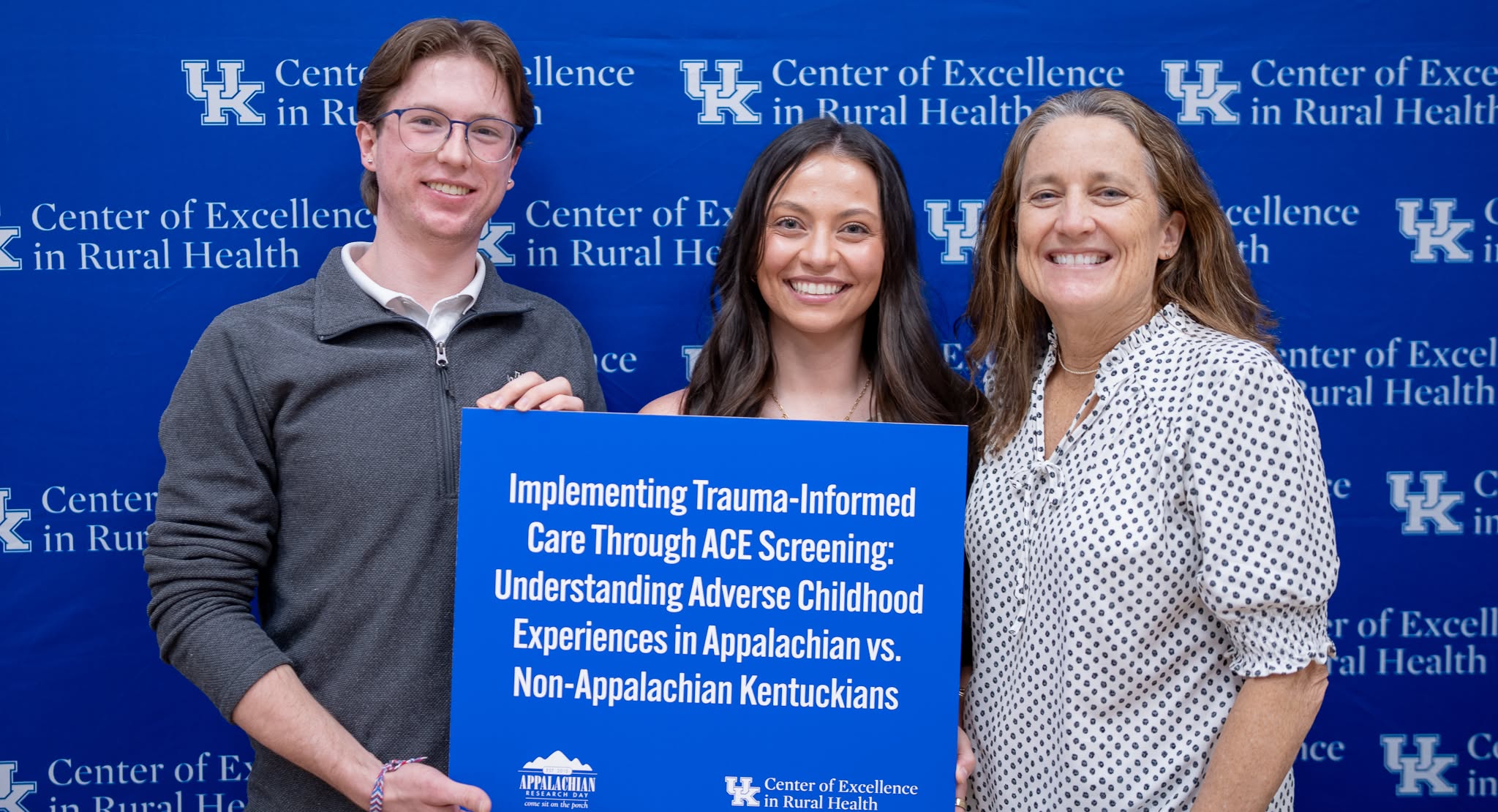 3 people pose in front of a blue background holding a poster that says "Implementing Trauma Informed Care through ACE Screening: Understanding Adverse Childhood Experiences in Appalachia vs Non-Appalachian Kentuckians