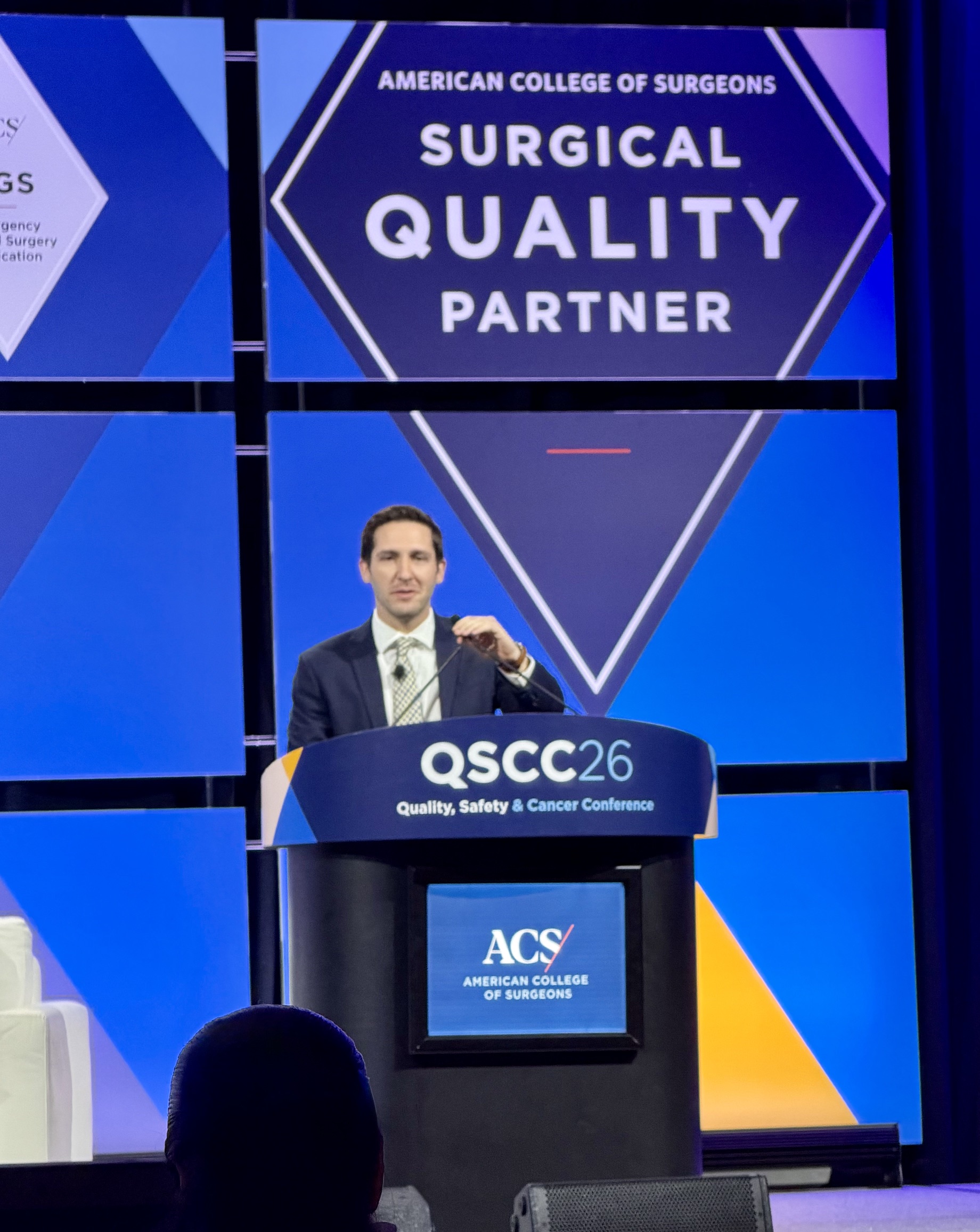 Dr. Jacob Hubbuch, in blue blazer and tie, stands behind podium labeled QSCC (stands for Quality, Safety, and Cancer Conference) while projected behind him is a blue diamond reading "American College of Surgeons Surgical Quality Partner."