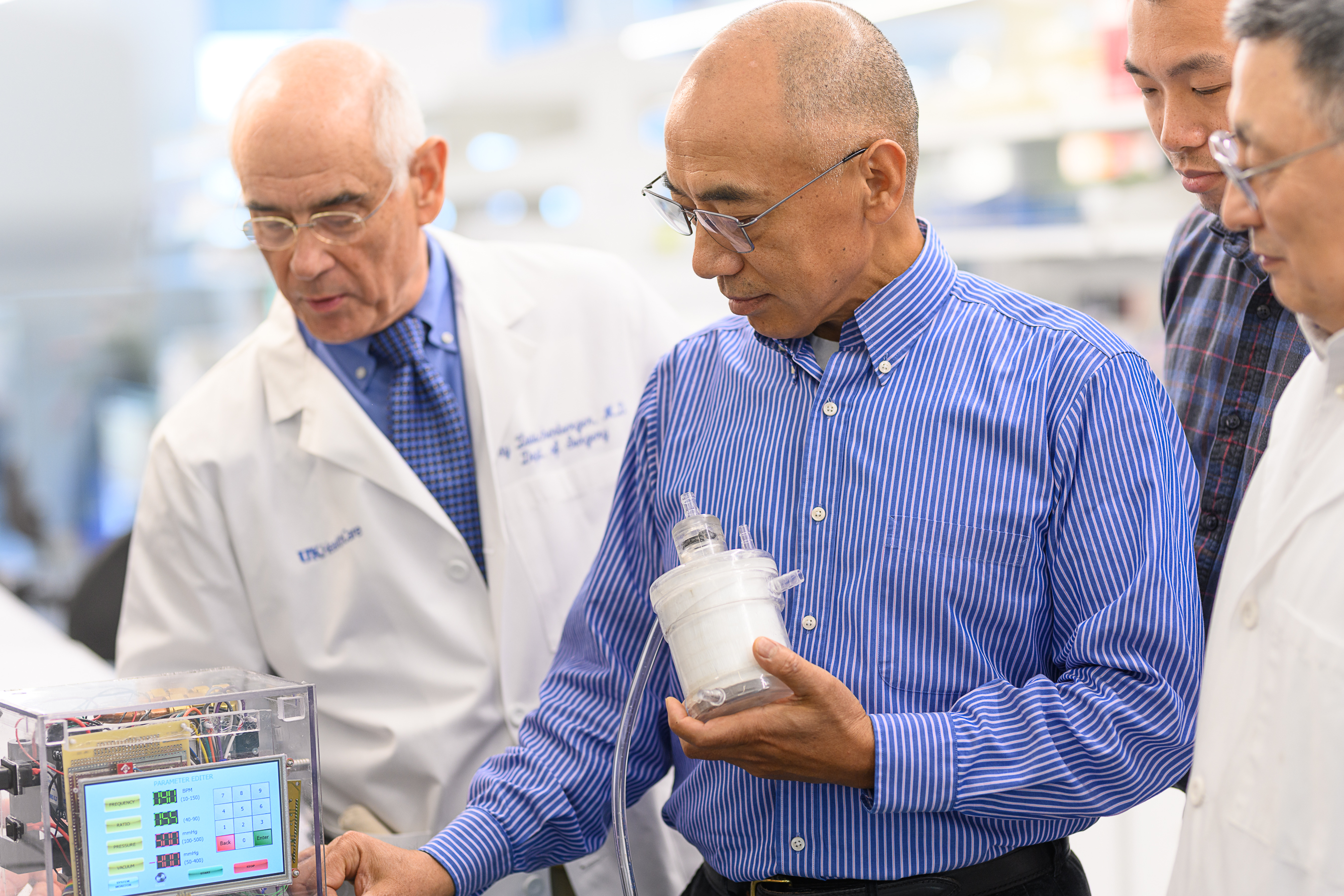 Dr. Joseph Zwischenberger, an older doctor dressed in white lab coat, looks at a clear box with wires and electronic displays. Next to him is  Dr. Dongfang Wang in a blue button down shirt demonstrates the function of a white medical assist device he holds in his hand that is wired to the box