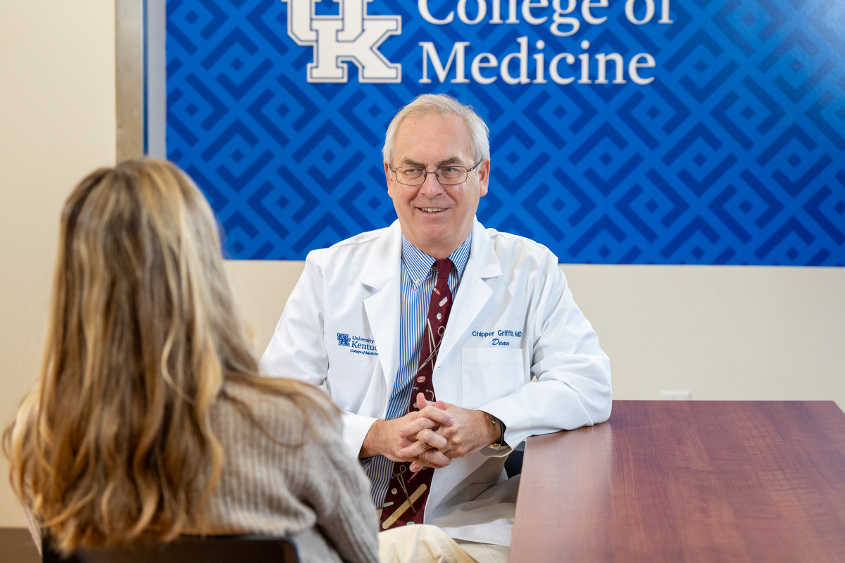 From left: The back of a woman's head as she sits and talks to  (right) Dean Griffith, wearing a white coat, smiling