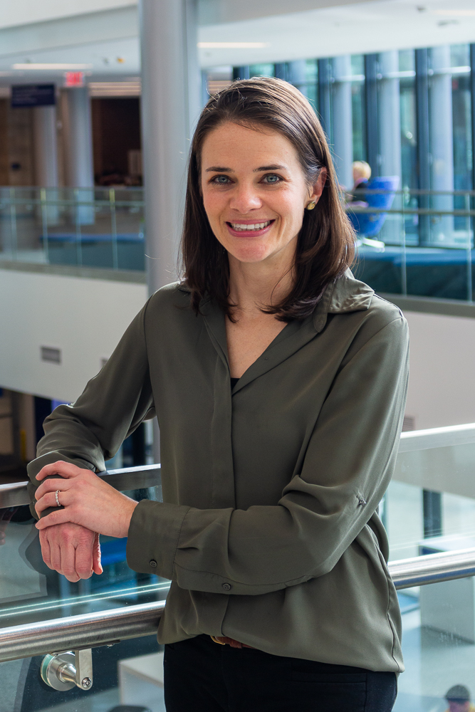 Woman standing on indoor balcony with arms folded in long-sleeve green dark green blouse with shoulder-length brown hair, smiling