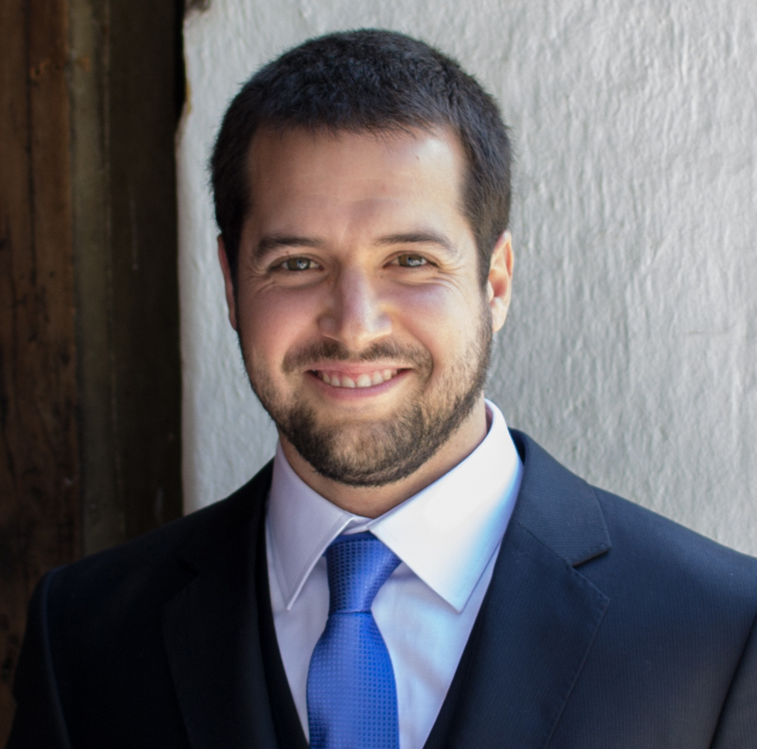 Man with short brown hair in black suit, white shirt and blue tie on white background