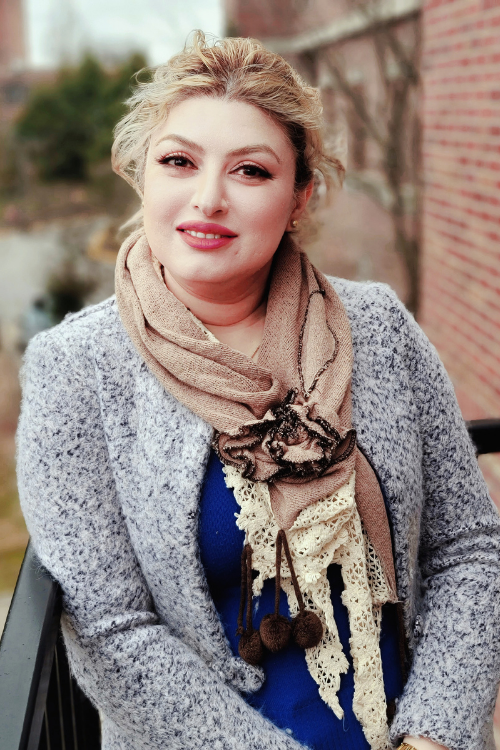 woman with blonde hair, gray sweater, tan scarf, and blue blouse, standing outside in front of brick building