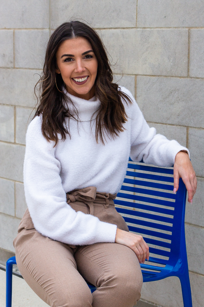 Woman in white turtleneck sweater sitting outside on blue bench in brown pants, smiling