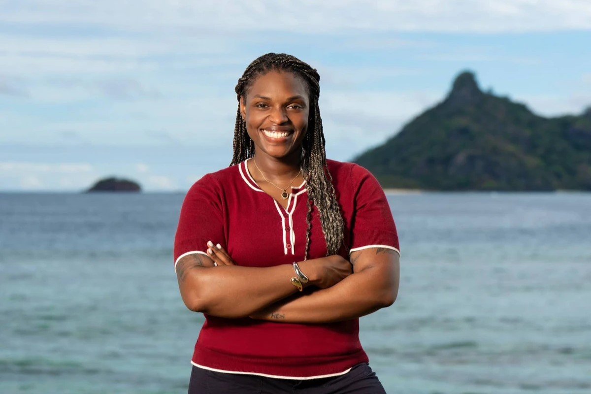 Sharonda Cox stands smiling, in a red shirt and black trousers stands with arms crossed on a tropical beach, with turquoise water, green islands, and a bright sky behind her.