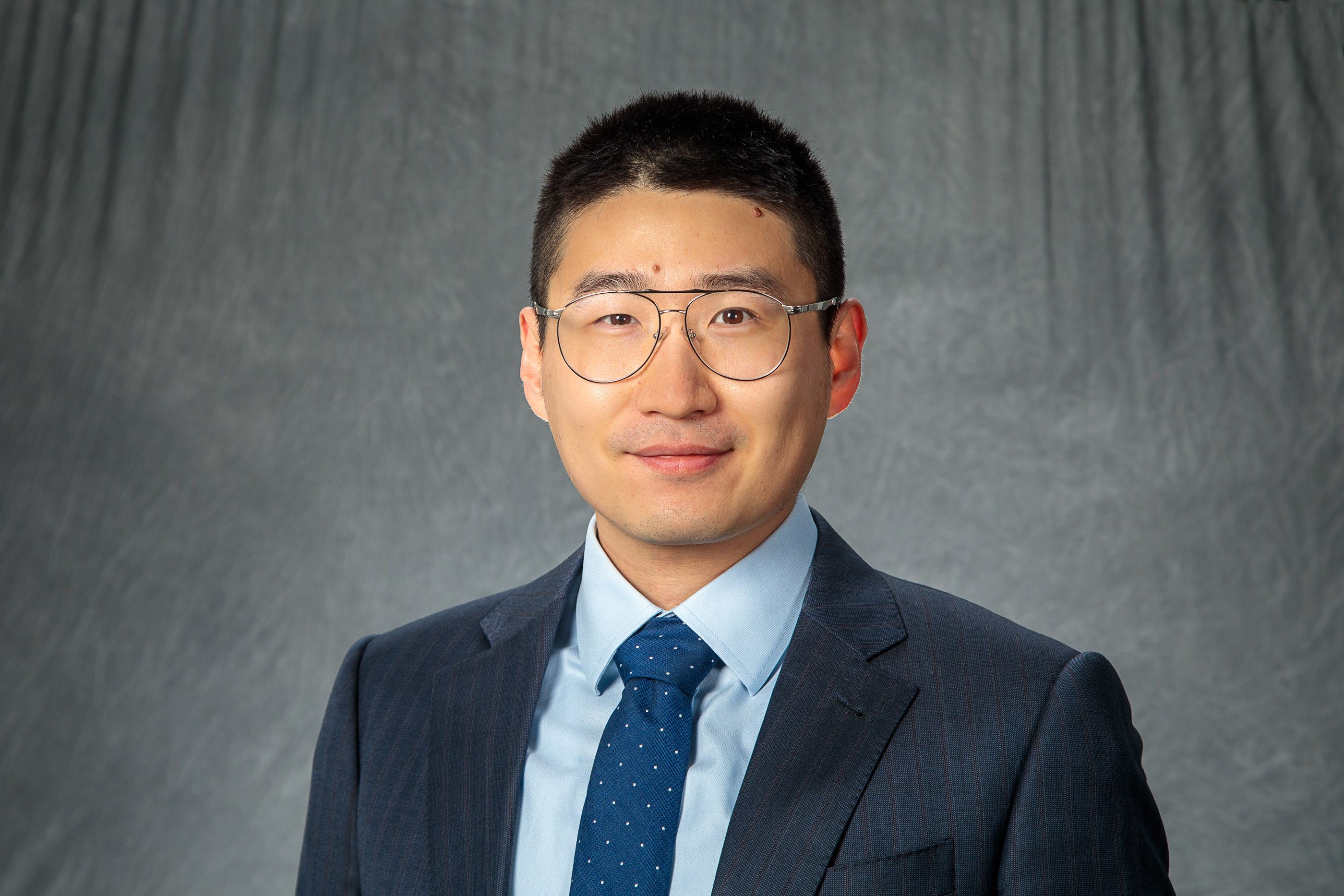 Photo of Man with short black hair, in a black suite, light blue shirt and blue tie on a gray background