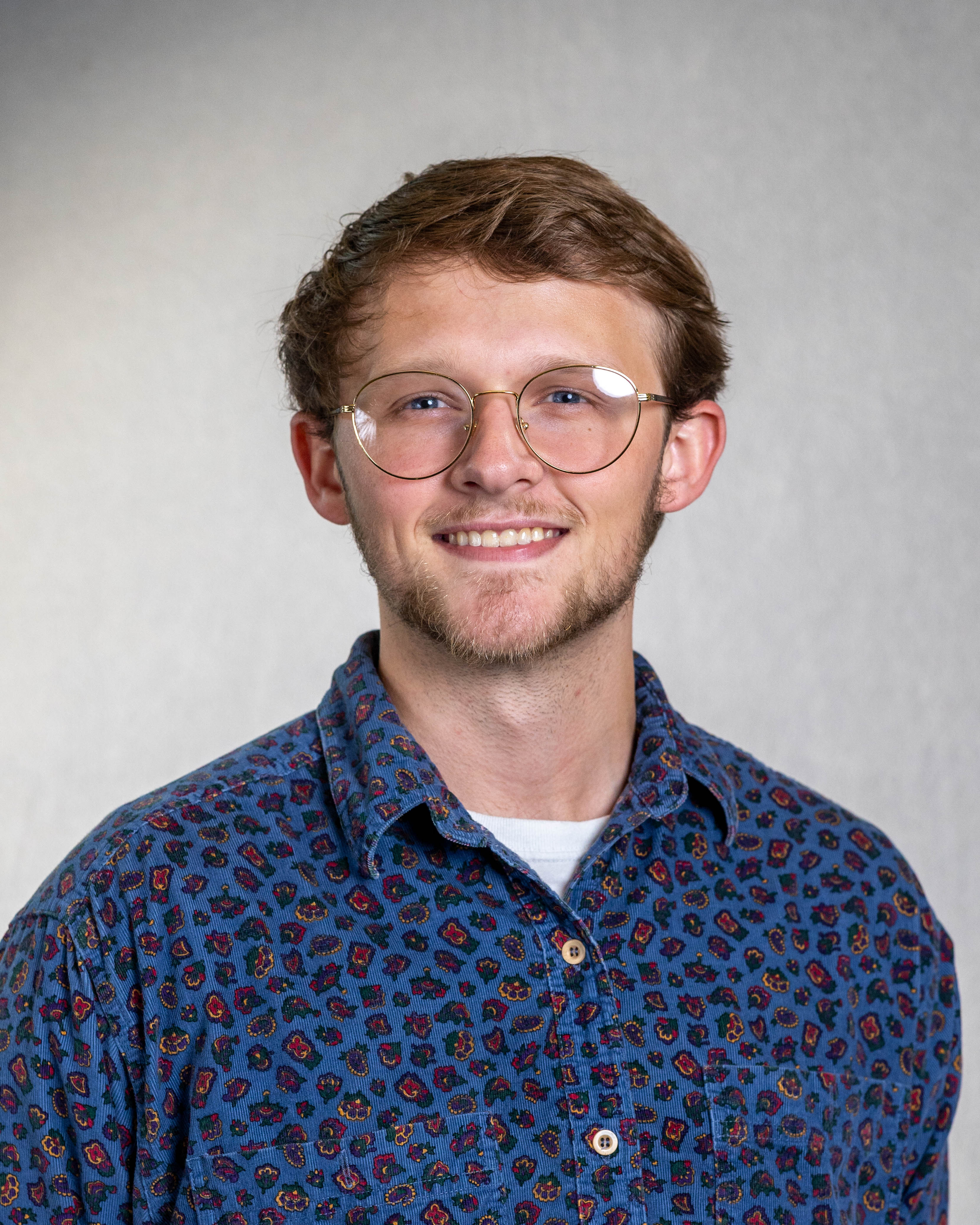 Young Caucasian man with glasses and blue button down shirt with brown hair smiling on gray background