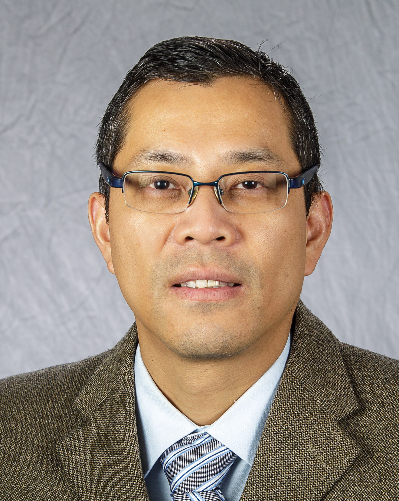 Man with black hair, glasses, brown suit, white shirt, striped tie on gray background