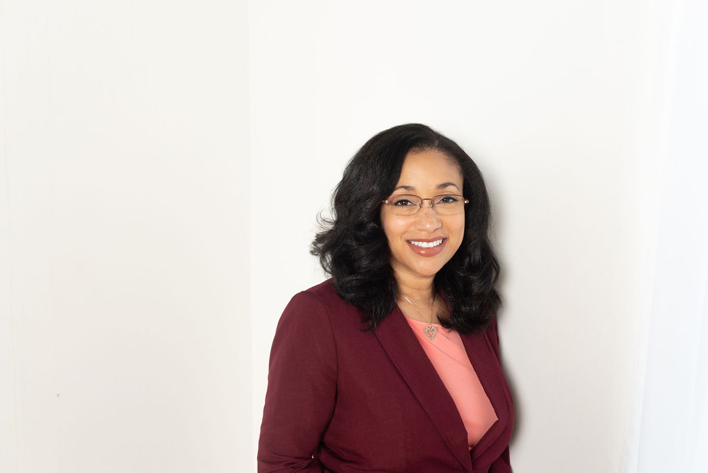 Woman with long dark hair in maroon blazer and pink blouse smiling on beige background