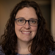 close up photo of woman with long curly dark hair and glasses smiling on brown background