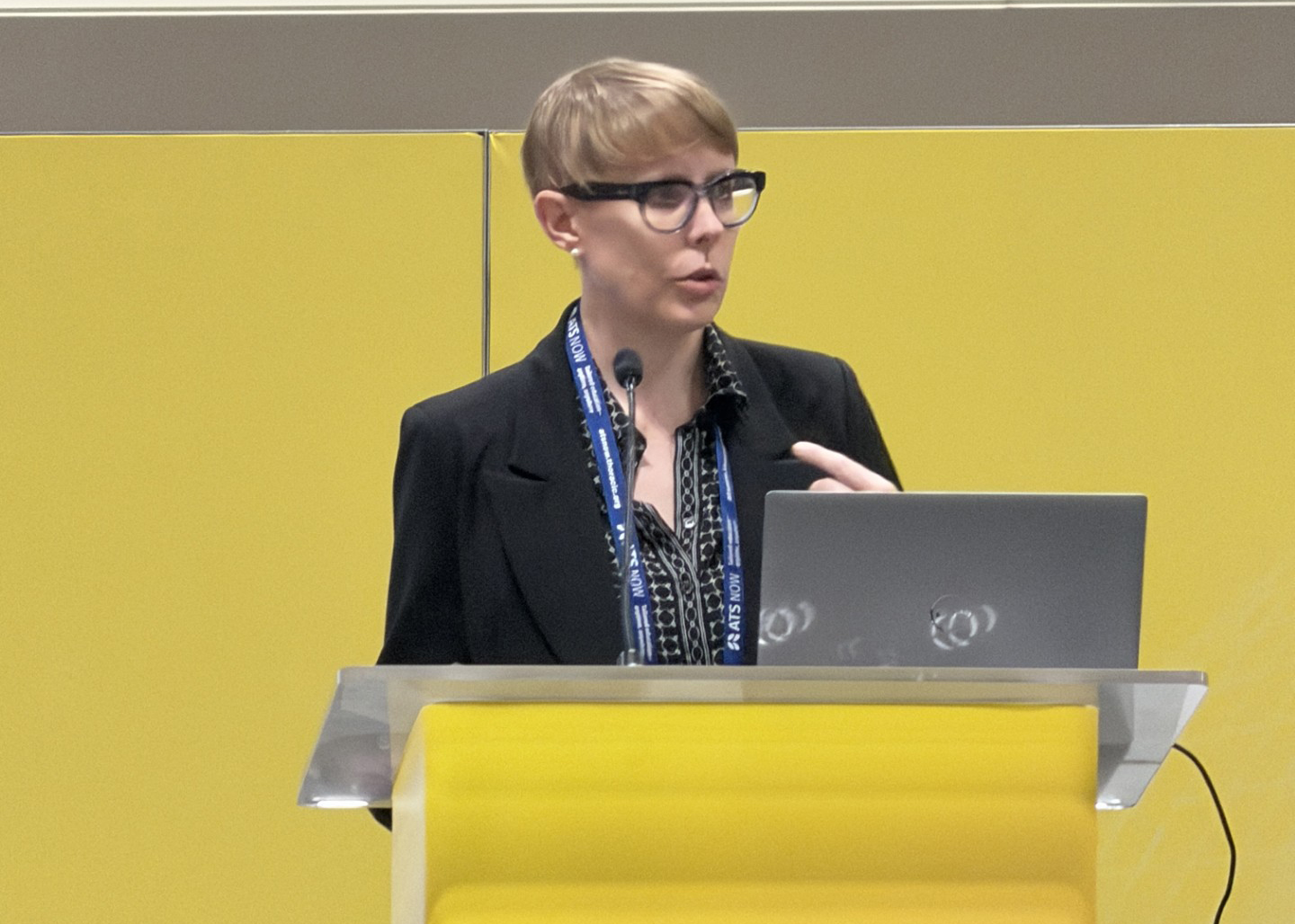 A short-haired speaker in glasses gestures behind a yellow conference podium with a laptop and microphone, presenting in a focused, professional setting.