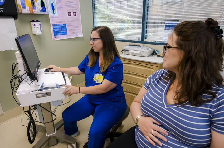 A health care professional in blue scrubs uses a computer beside a seated pregnant patient in an exam room.