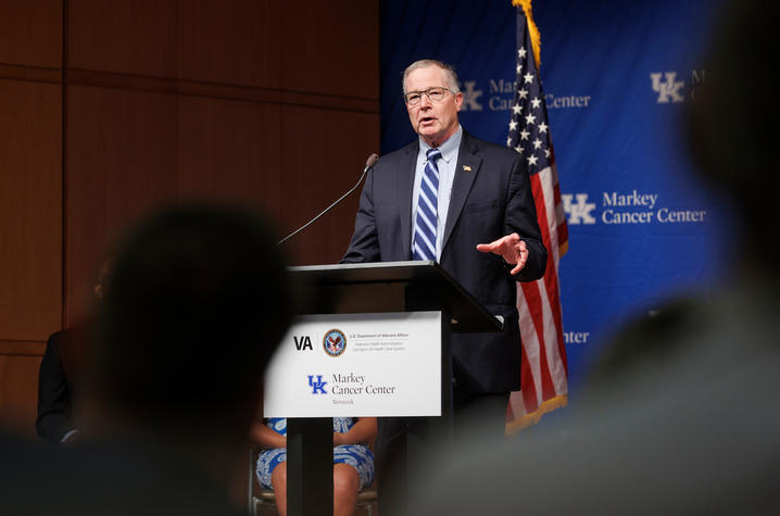 A person wearing glasses, a dark suit and striped blue tie speaks at a podium during a formal event. An American flag and a blue backdrop displaying the UK Markey Cancer Center logo are behind the speaker.