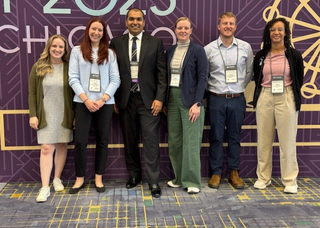 Six conference attendees wearing name badges pose together before a purple-and-gold 2023 event backdrop, smiling in a friendly, welcoming group photo.