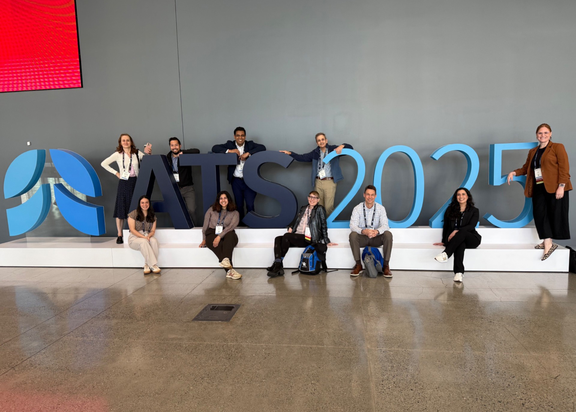 A diverse group of eight people pose around oversized blue “ATS 2025” lettering at a conference venue, smiling in a relaxed, welcoming atmosphere.