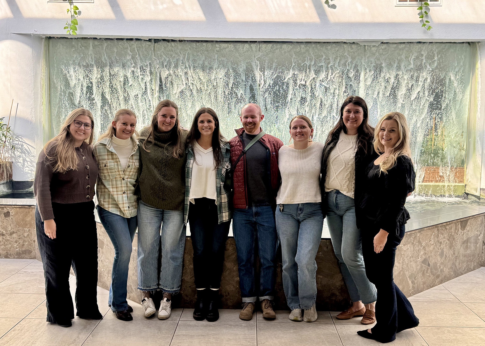 Eight smiling adults pose arm-in-arm in front of a large indoor waterfall, wearing casual outfits in a bright atrium; the group photo feels warm and celebratory.