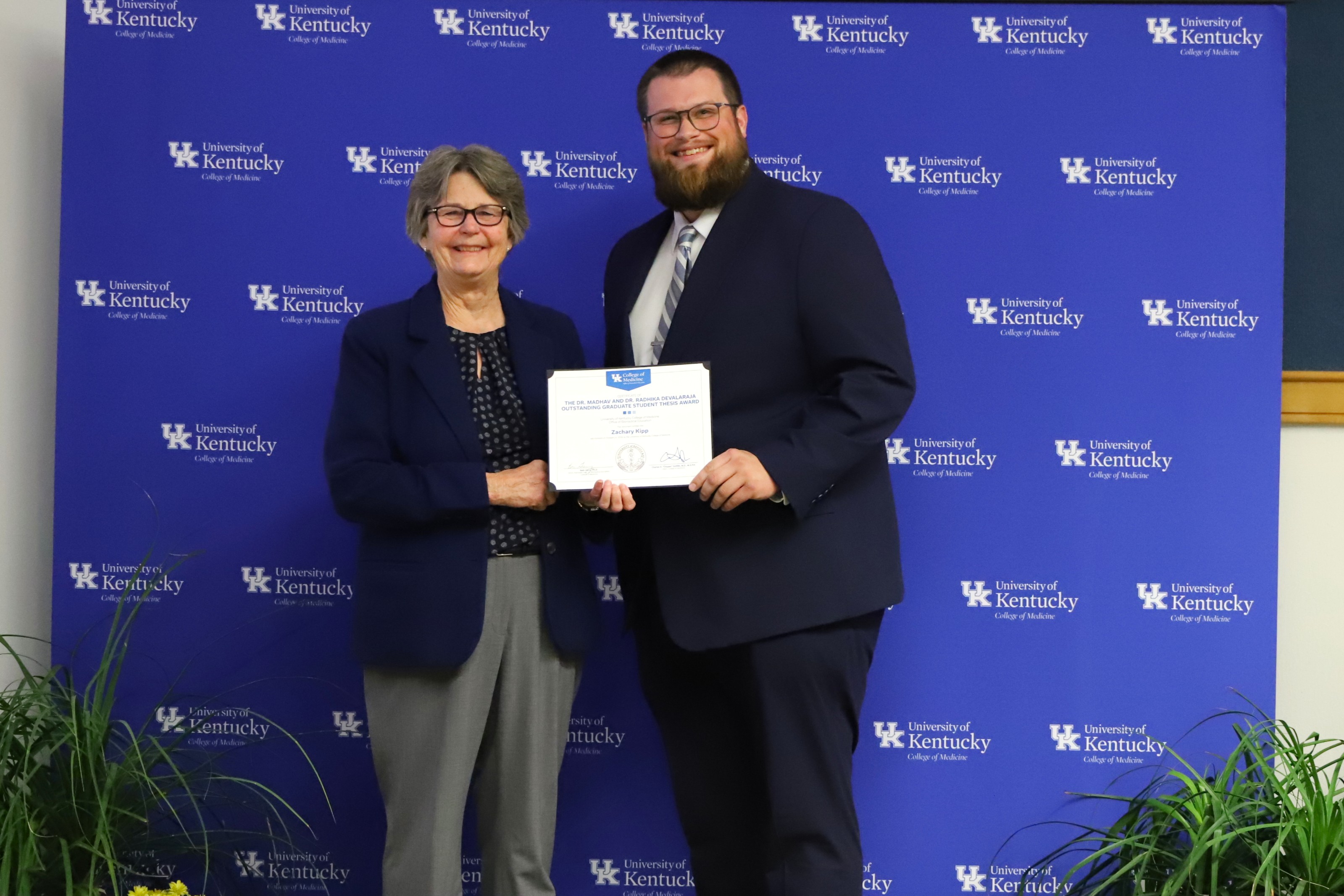 Dr. Garvy and Zachary Kipp pose in front of a UK College of Medicine backdrop, smiling. He holds a certificate. 