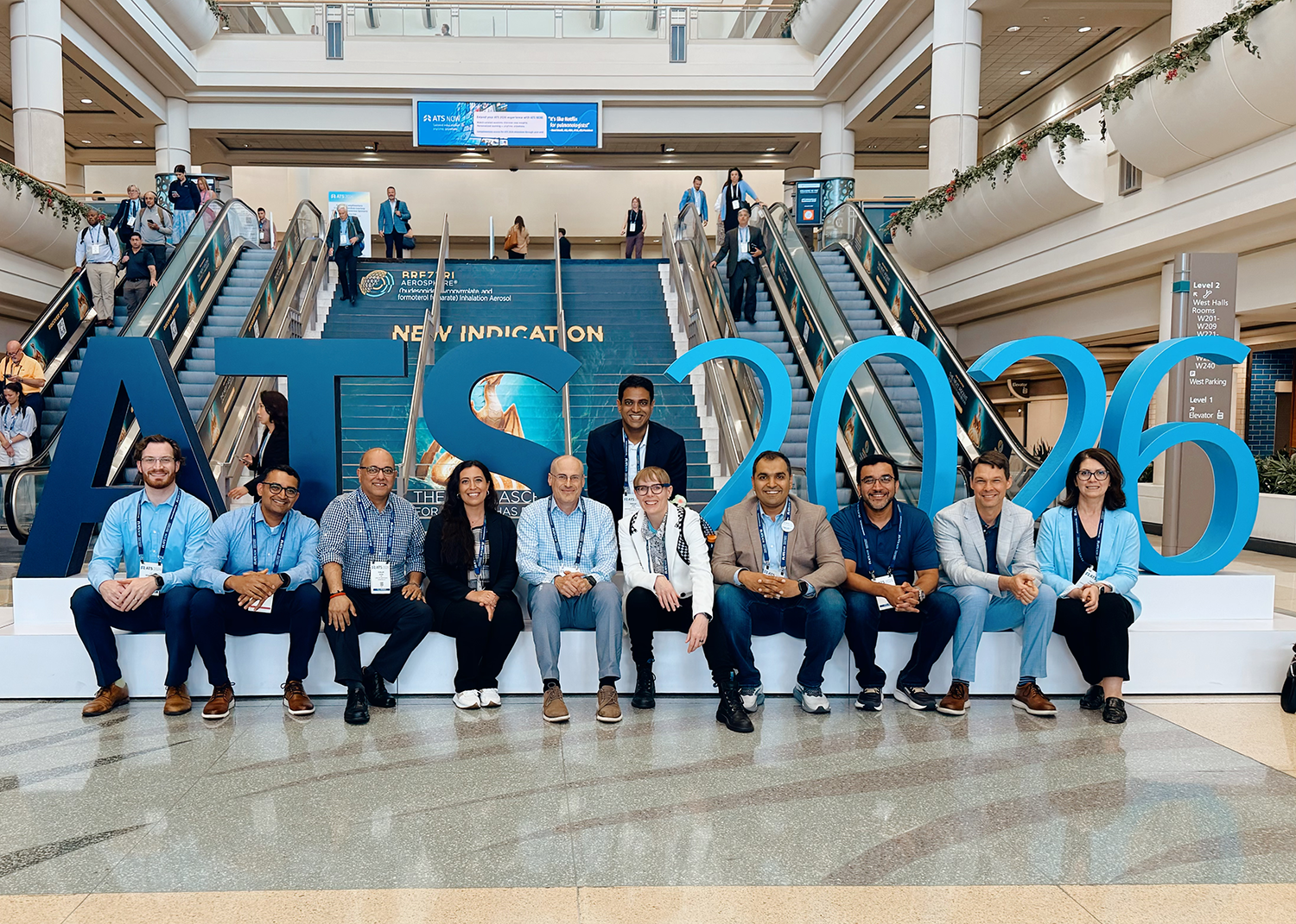 A diverse conference group poses beneath a large blue “ATS 2026” display in a bright convention hall, conveying a welcoming, upbeat event atmosphere.