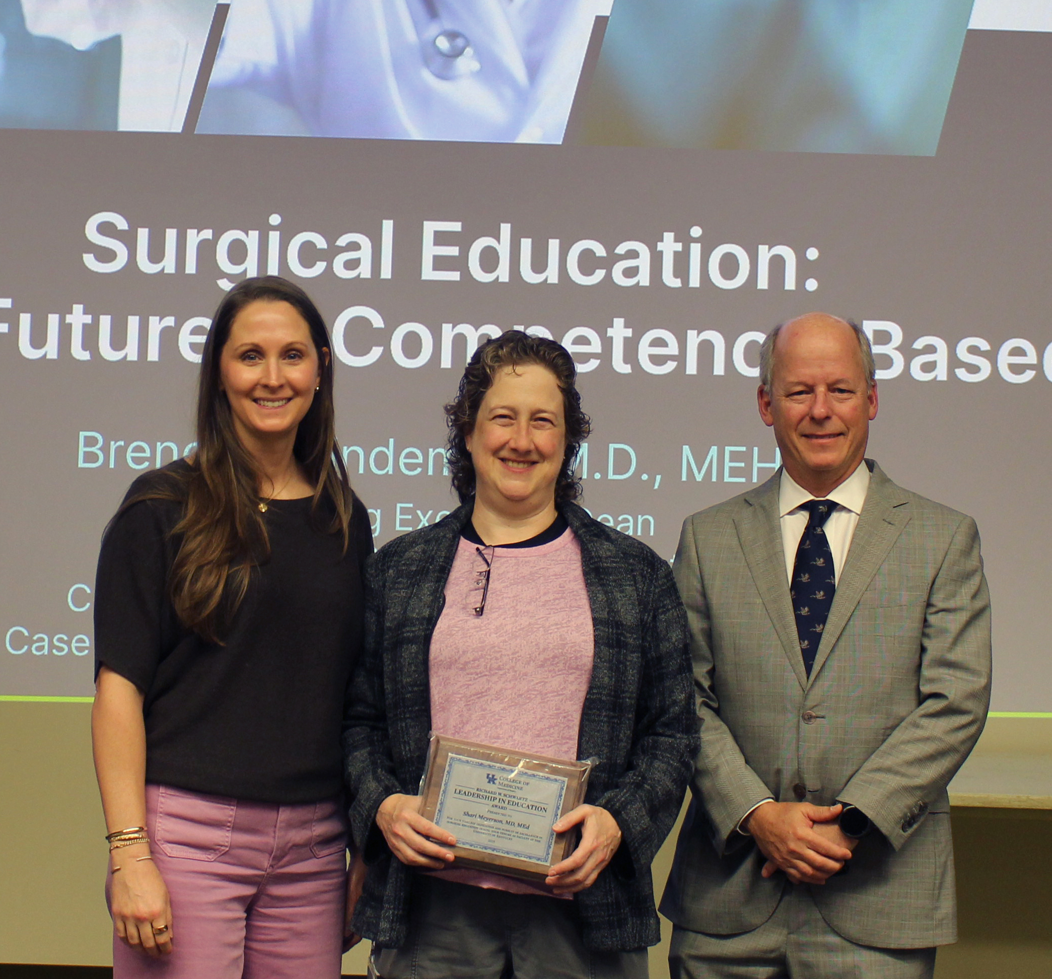 Doctor Shari Meyerson, center, holds a Schwartz plaque in business dress. On her left is Doctor Melissa Ranney Newcomb, Vice Chair for Education, in casual pants and blue sweater. On her right is doctor William Inabnet, Chair of the UK Department of Surgery, in a light gray business suit. 